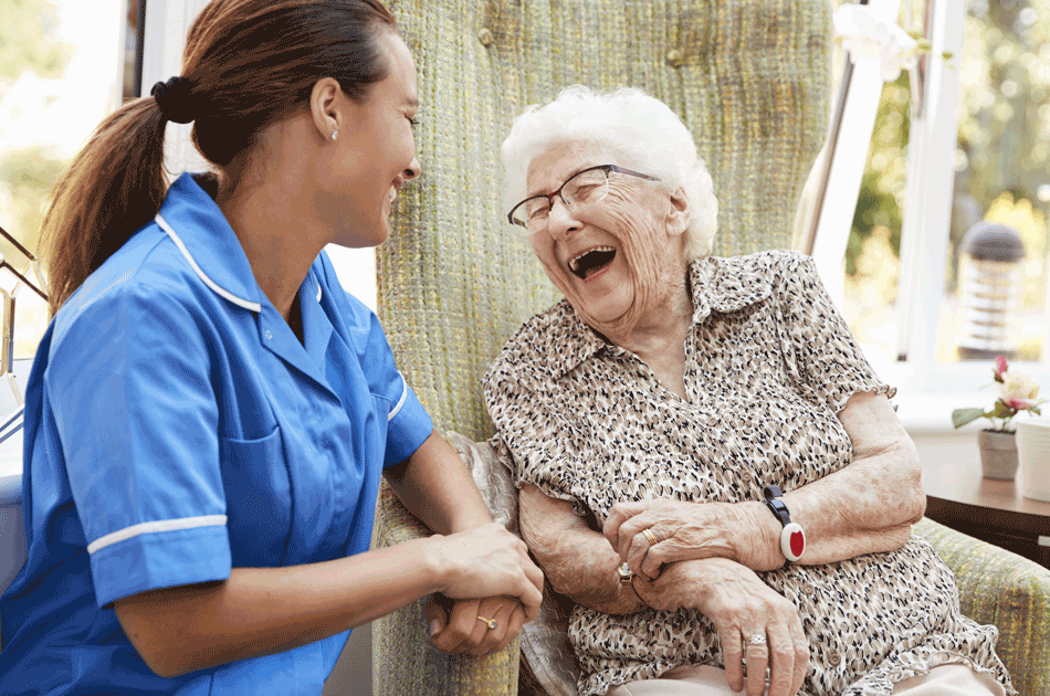 Eine Pflegegkraft unterhält sich mit einer älteren Dame, welche im Sessel sitzt und herzlich lacht.