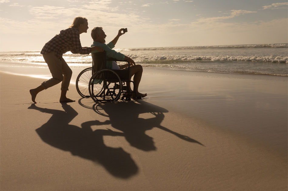 Ein Paar am abendlichen Strand. Er sitzt im Rollstuhl und Sie schiebt ihn.