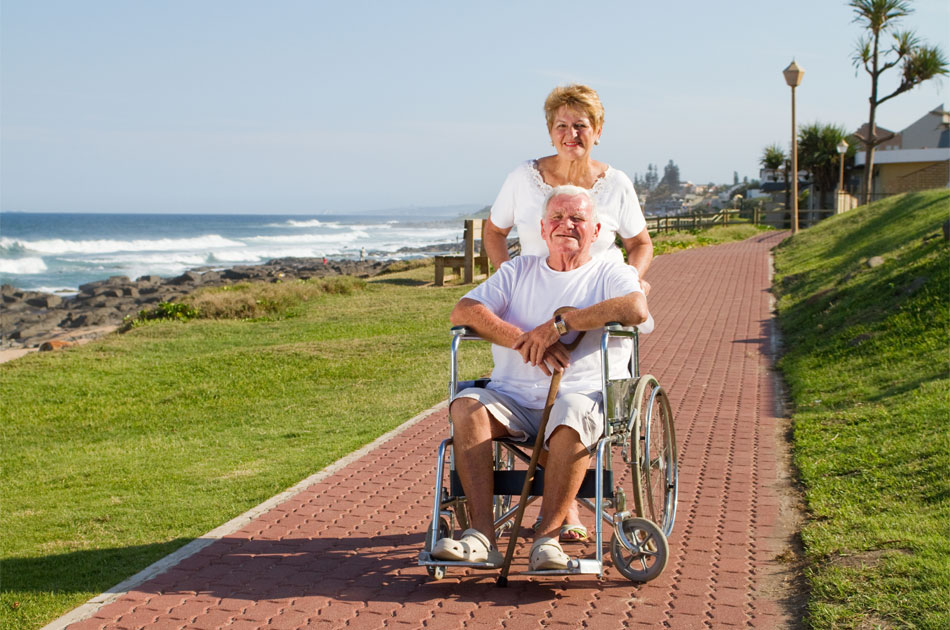 Eine Urlaubsbetreuerin schiebt einen Pflegebedürftigen in seinem Rollstuhl an der Küste entlang.