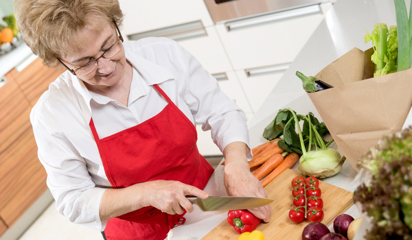 Eine Seniorin kocht gesund mit Gemüse und Möhren, welche sie sich auf einem Brett klein schneidet.